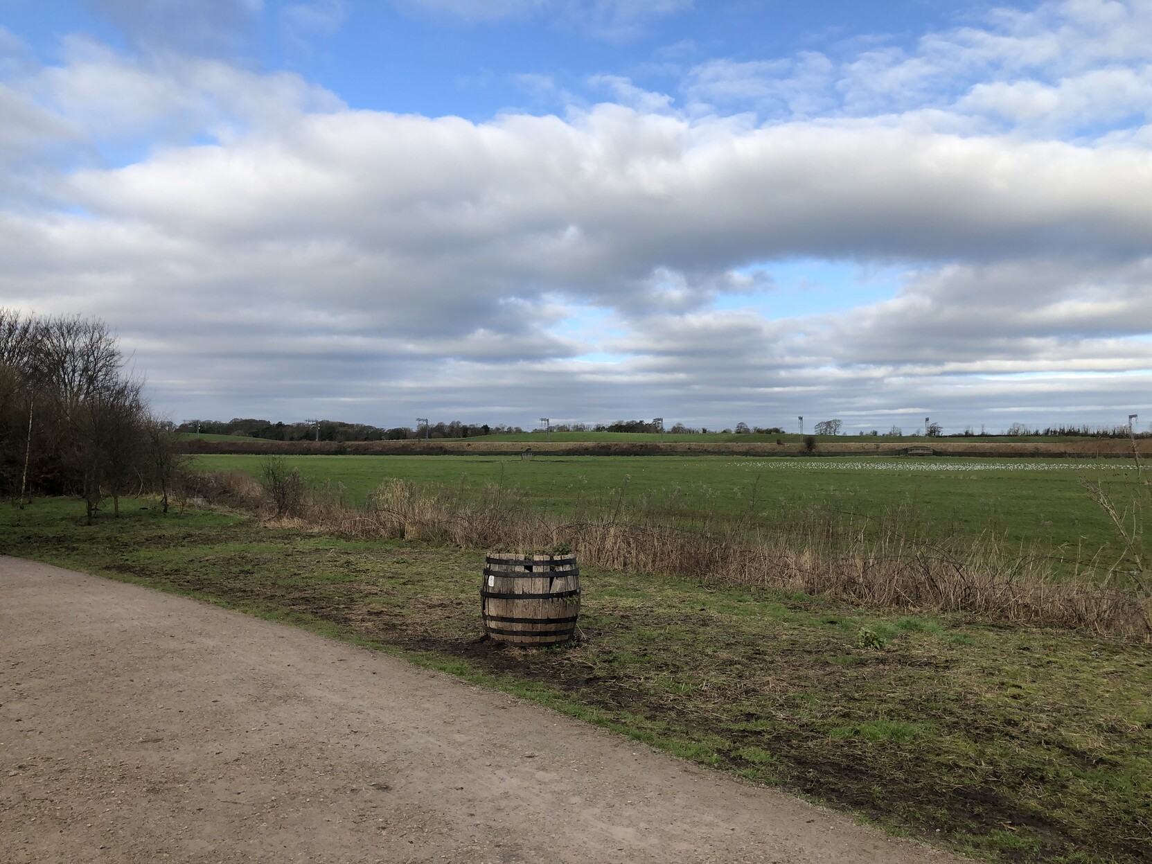 A view of an ordinary local field, with a railway embankment running all the way across the view, and more fields in the background. A dilapidated wooden barrel is in the middle foreground and there are some weeds growing out of the top.   

The sky is covered in low cloud, but there are still large patches of blue visible.  Despite the blue, there is no evidence of what one would call sunshine.

The clouds stretch out into the distance, and appear to have been arranged into neat rows across the view.