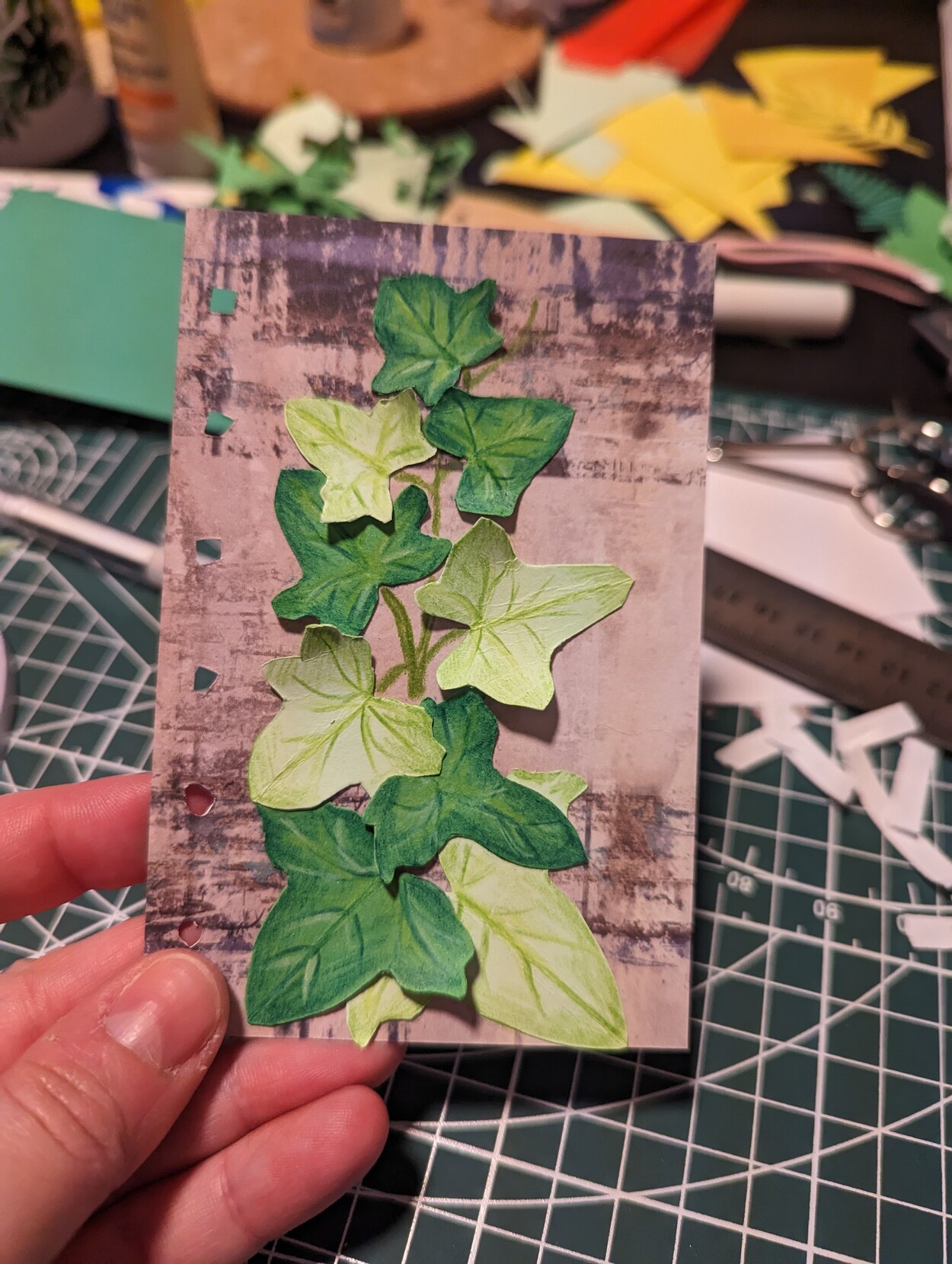 An A6 sized journal page consisting of a grungy grey background paper and a set of cut out green paper leaves resembling an ivy vine. 