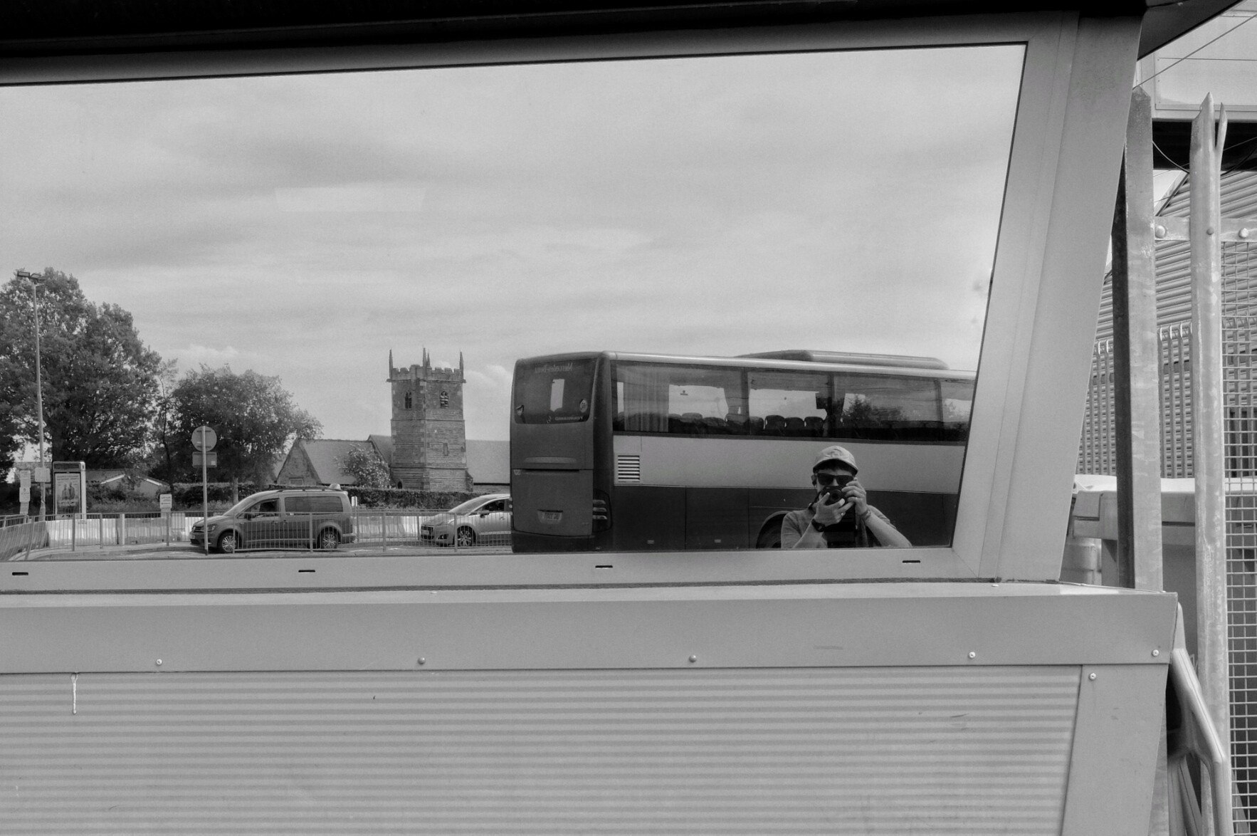 A horizontal black and white photograph of a reflective window by the side of a metal building. The mirror takes most of the frame, reflecting a quiet street scene with cars parked, the square spire of a church, trees in bloom. At the forefront a bus is parked and me the photographer can be seen creating the image on the left hand corner.