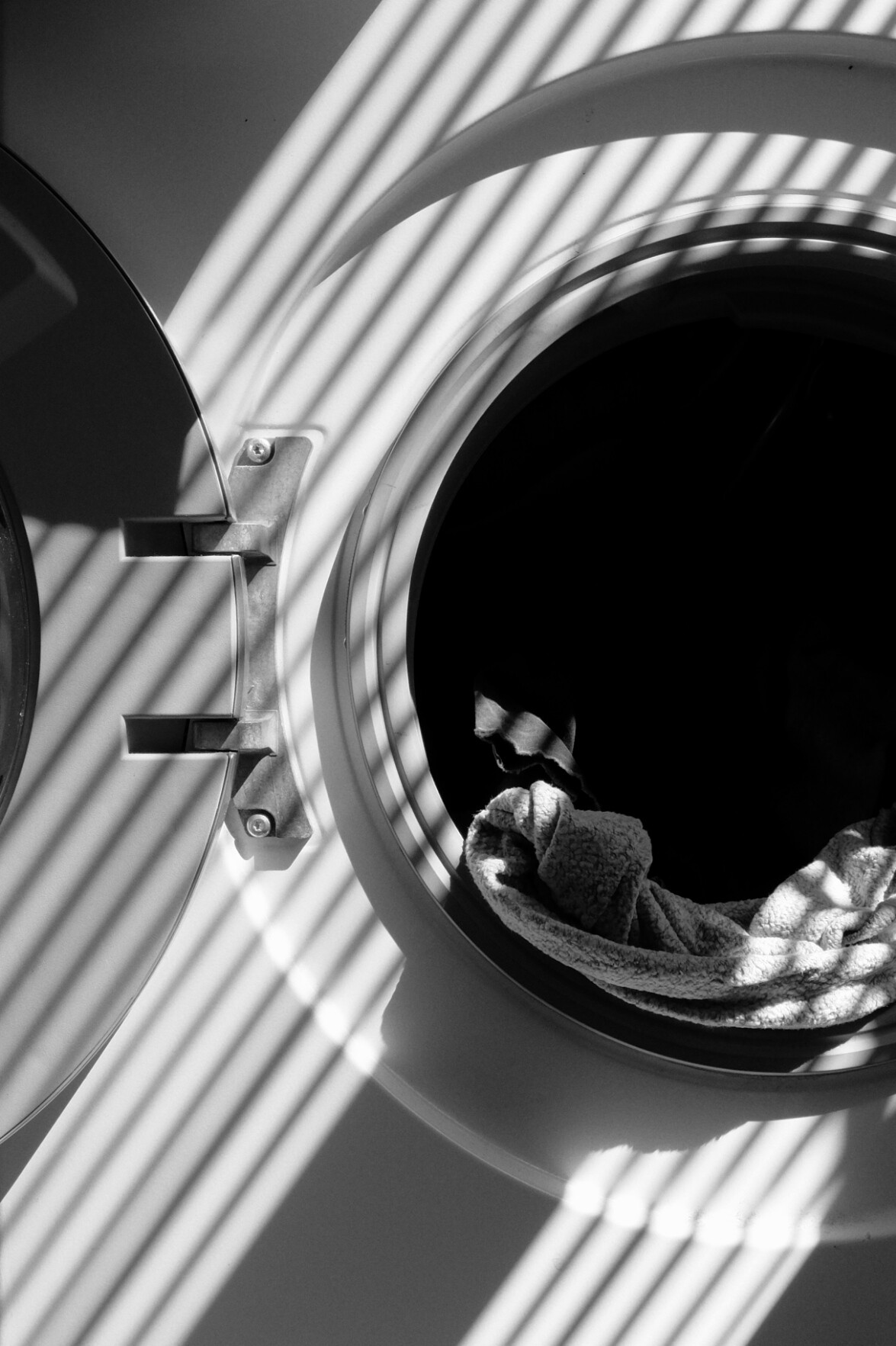 A black and white vertical photograph of a washing machine. The image is a close up of the open door of the washing machine with neither the opening or the door fully in frame. There is laundry in the washing machine but only the clothes poking out are visible. The rest falls into darkness. Streaks of diagonal light and shadow fall across the scene.