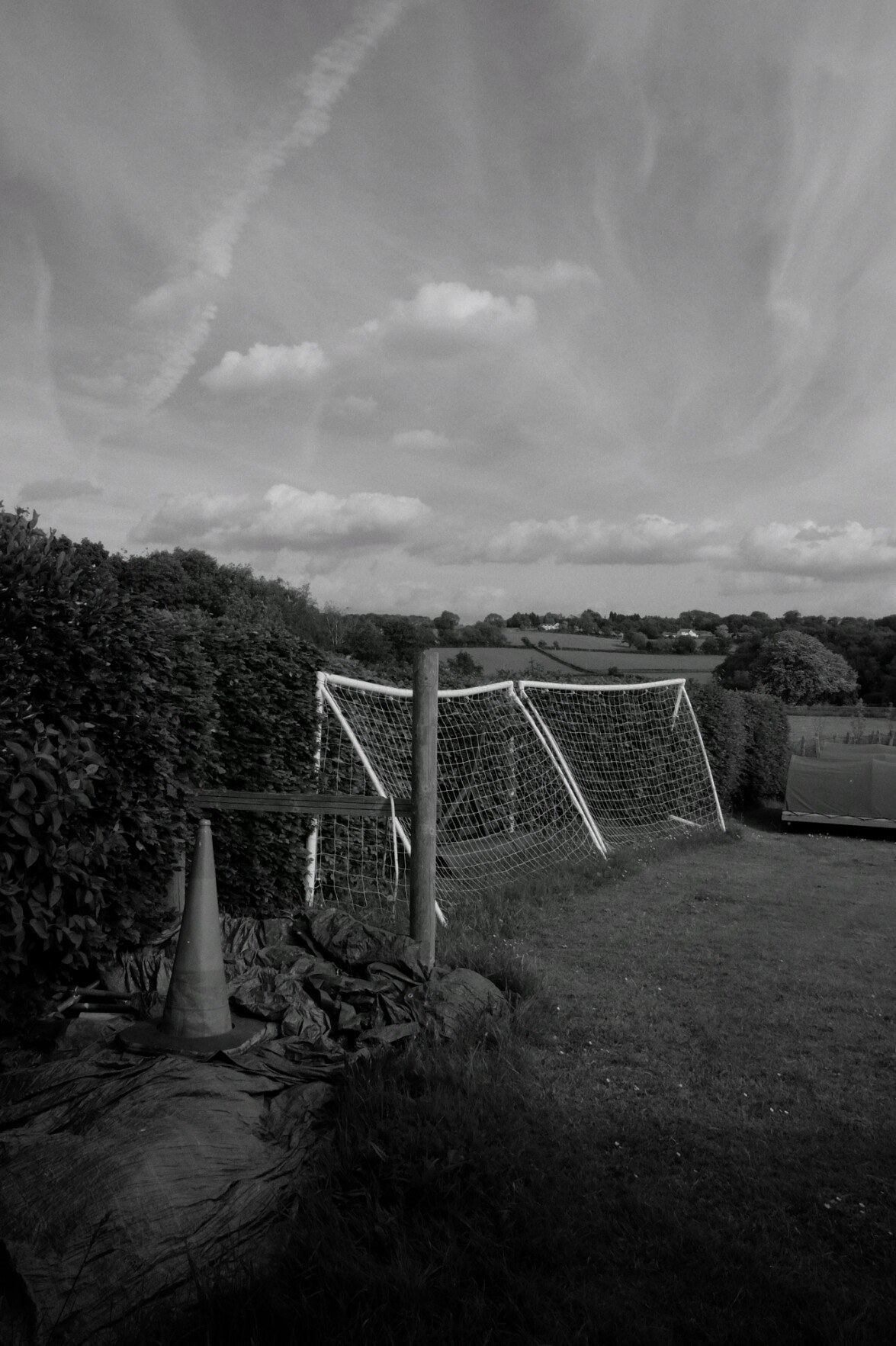 A black and white vertical photograph of a field with a two side by side football cages turned the wrong way resting against a hedge. Some cones are at the side with other sport equipment. In the distance more fields. Trails of clouds in the sky.