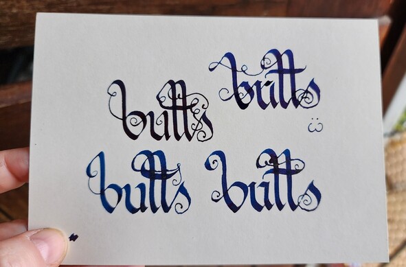 Flat top down view of a piece of paper with the word 'butts' calligraphied on it four times in dark ink, in the same style, with long, swirly ascenders and lots of curls