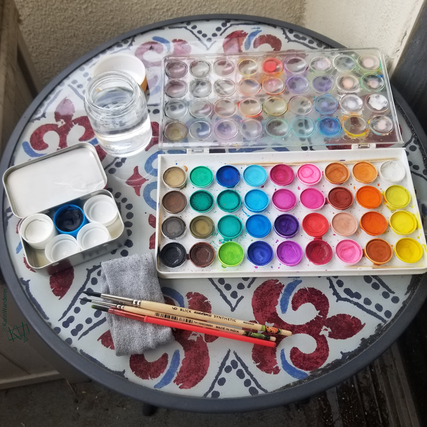 Outside art kit arranged on a small, round table. Glass of water with lid, cloth, 3 paintbrushes, many-colored watercolor palette, & a black & white gouache palette.