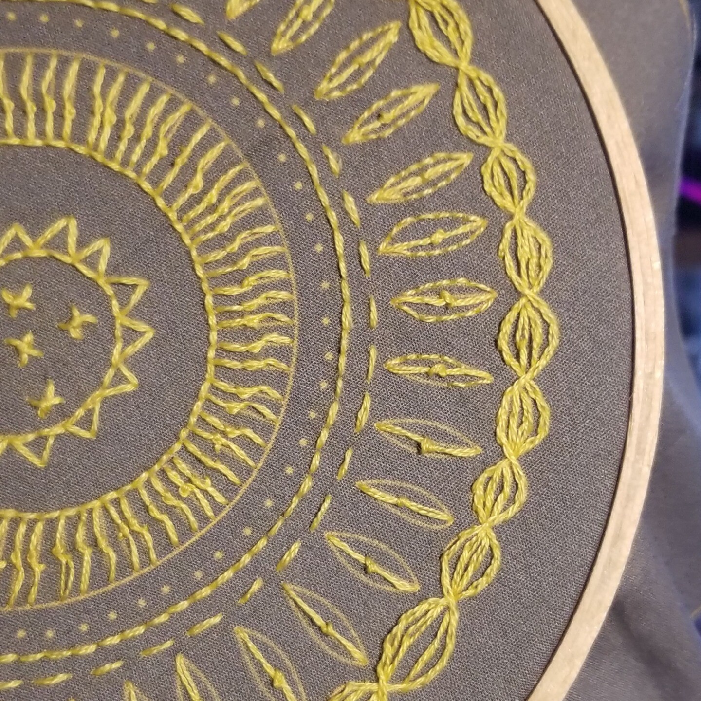 Almost completed embroidery of a sunflower mandala in yellow thread on a grey background.