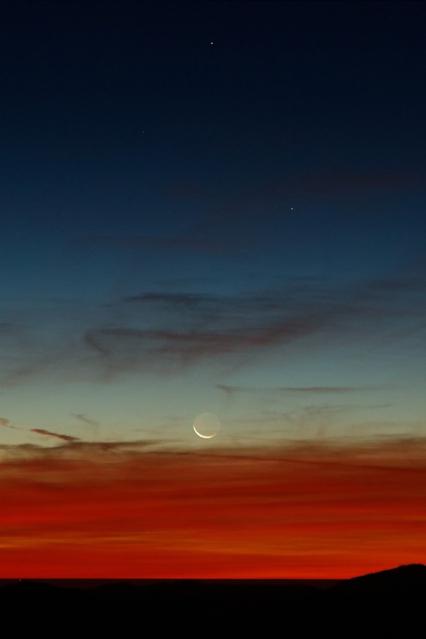 A fiery red sunset on wispy clouds at the horizon with a darkening sky above and a few stars. Just above where the clouds end is the moon, with a  small crescent and visible outline of the whole thing.