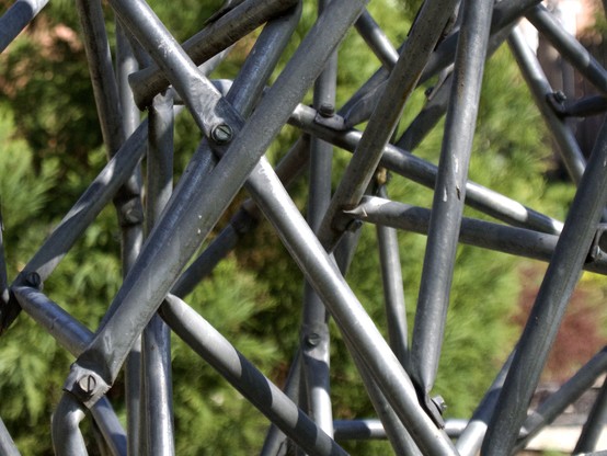 Closeup of a metal sculpture consisting of interlocking aluminum tubes, sunlit trees out of focus in the background