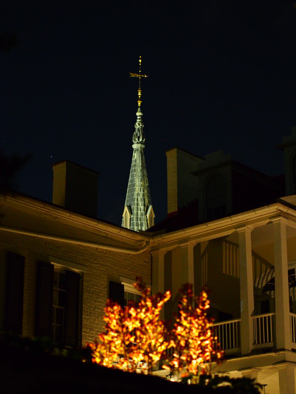 A brightly lit, weathered, slate-tiled church steeple rises into the black night sky behind a yellow-tinted house with a brightly lit bush out of focus in the foreground