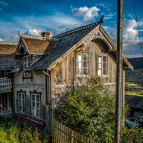 A more than a hundred-year-old abandoned two-storey residential building in the evening sun. Two windows in the second bay, in the gable wall, against the sun coming in from the right. Three windows in the shadow on the left. The house has a slate roof and joinery decorations along the ridge. But everything is in total disrepair, gray and sad. A stately house - long ago, with a beautiful patina. In the foreground, an equally unpainted picket fence slanting towards us in front of a tree with green leaves close to the house which reaches right up to the windows on the gable wall.
The evening sky above the house is deep blue with small white clouds.