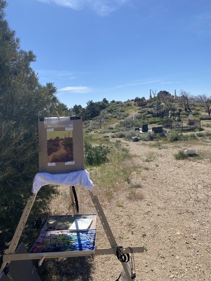 An outdoor easel with small work-in-progress painting on, in a tiny shade by a tree 