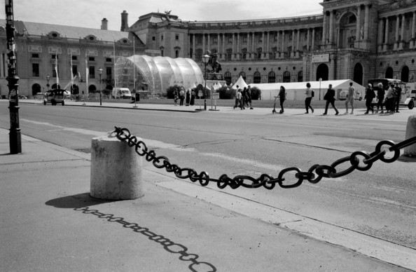 Black and white picture of a big iron chain next to the street casting a shadow, in the background tourists walking in front of the Hofburg.