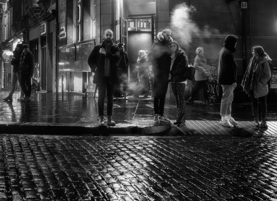 A black and white photo from the series by Lynn Henni called Edinburgh Nights, giving her perspective on Edinburgh night life.

A cold, rainy and damp looking street corner, as different people decide what to do next, partially enveloped in a light cloud of moisture. Edinburgh,| Scotland.

Photo by and copyright of Lynn Henni.