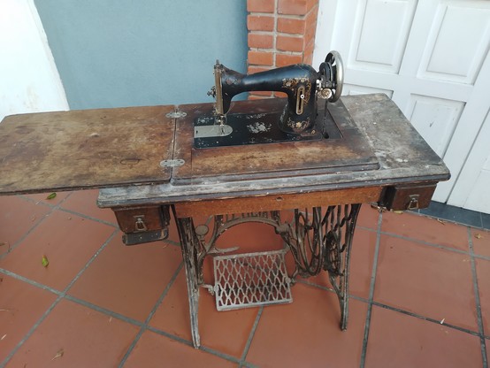 Foto de una mesa con máquina de coser antigua a pedal, sucia pero entera, abierta, de hierro medio despintado, hasta tiene la aguja de coser.
