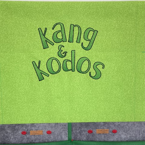 Green minimalist quilt block on a design wall; the block has the names Kang & Kodos in a fun green ombré font with a gray section below that resembles the alien’s collars 