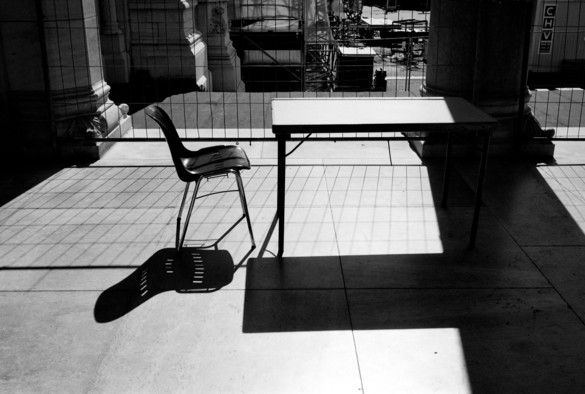 The image shows a single plastic chair and a rectangular table positioned on a sunlit terrace. The strong sunlight casts distinct shadows of the chair, table on the ground. In the background, there is a wire fence and construction equipment, indicating an ongoing construction site. The scene emphasizes the stark contrast between light and shadow.