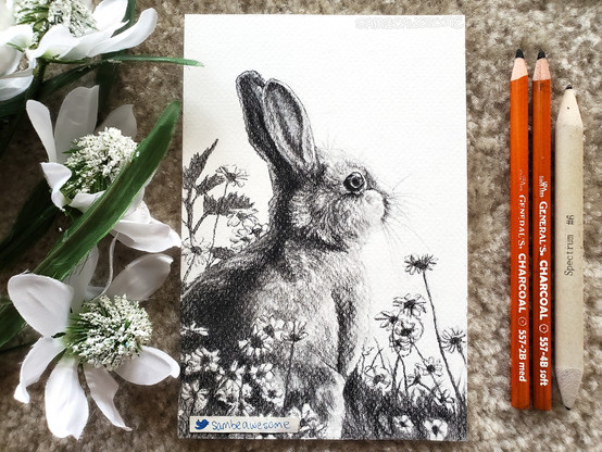 A photo of a charcoal drawing of a rabbit that's surrounded by flowers and grass. Faux flowers are next to the artwork for flair, along with the pencils used to make the artwork.