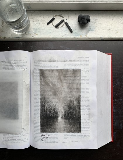 Photo of a charcoal landscape sketch in the middle of a gessoed law dictionary page. Printed words on the page are partially visible beneath the gesso and charcoal. On the windowsill at the top of the photo is a glass of water, three pieces of charcoal, and an eraser.