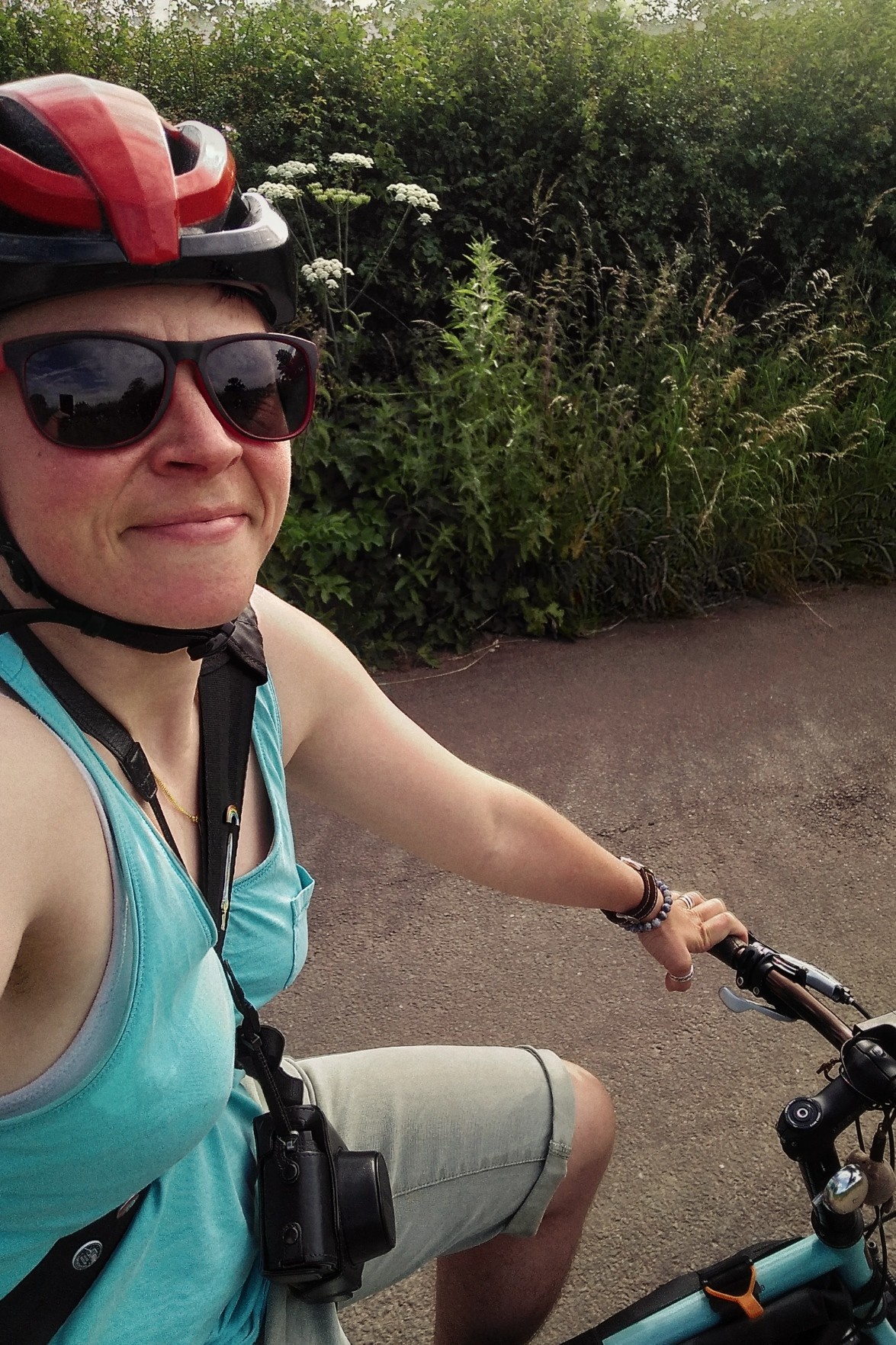 A vertical colour photograph of me (a white woman) smiling whilst cycling in shorts and t-shirt with sunglasses on.