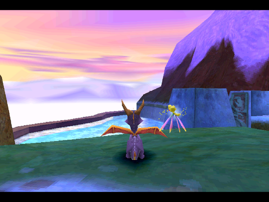 A screenshot from the level colossus from Spyro 2. You can see some dark grass above a frozen lake, and a pink and yellow sky with white mountains in the distance.