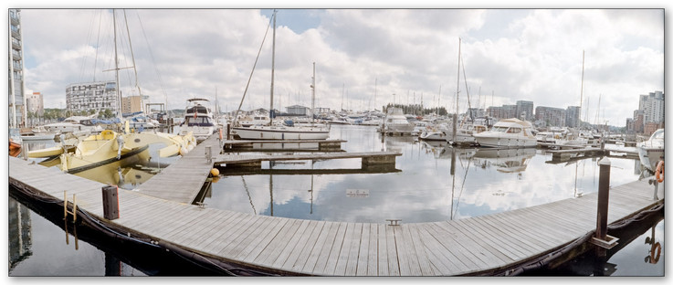A colour panoramic photograph of Ipswich marina. Floating pontoons and walkways with yachts and motor boats moored. A yellow trimaran sits on the left side. Clouds and blue sky which are reflected in the flat water. Apartment buildings can be seen on either far edges.