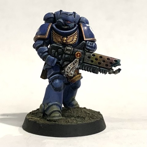 A Space Marine in dark blue power armor with giant shoulders, purity seals, and various gold adornments. They carry large flame-thrower with a rainbow heat bloom on the barrel's radiators. They face to the right of the photo, ready to fire. They stand on rough brown and beige soil.