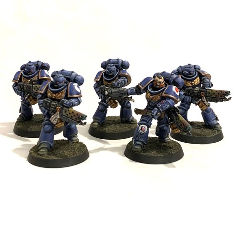 A squad of five Space Marines in dark blue power armor with giant shoulders, purity seals, and various gold adornments. They carry large flame-throwers with a rainbow heat bloom on the barrel's radiators. The Sergeant has his flame-thrower slung over his shoulder while he pulls out a long blade from a sheath on his hip. He has a pistol in his other hand. They all face to the right of the photo, ready to fire. They stand on rough brown and beige soil.