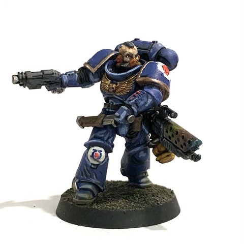A Space Marine in dark blue power armor with giant shoulders, purity seals, and various gold adornments. The Sergeant has his flame-thrower slung over his shoulder while he pulls out a long blade from a sheath on his hip. He has a pistol in his other hand. They face to the right of the photo, ready to attack. They stand on rough brown and beige soil.