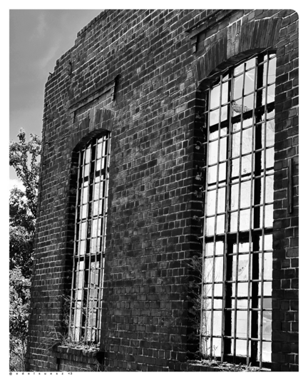 Schwarzweißfotografie: alte Industriebrache, freistehende Ziegelmauer einer Ruine, dunkle Steine mit hellen Fugen, mit zwei großen vergitterten Fensterfronten, im Hintergrund ein Stück eines Baumes und hellgrauer Himmel darüber  
