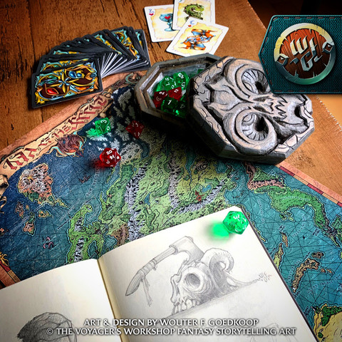 An open sketchbook with a pencil illustration of an axe embedded in a demon skull, surrounded by a fantasy map, cards, dice and their wooden box.