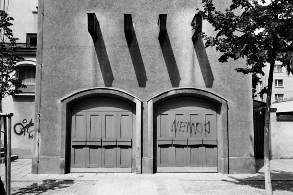 A black-and-white photo shows a building facade with two large, closed, arched doors. The doors have a paneled design and the word “NEMO” is graffitied on one of them. Above the doors, there are three box-like structures casting shadows on the wall. A tree is visible on the right side, and another building is partially visible in the background.