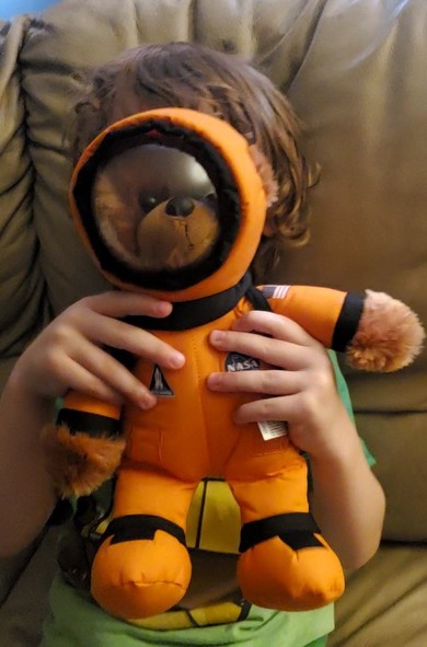 A young child holds a teddy bear dressed in an orange space suit in front of himself.