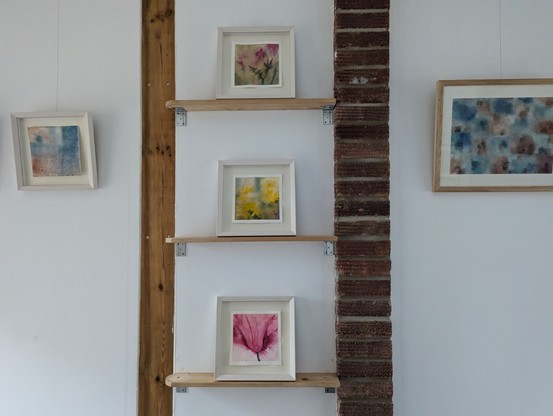 Photo of a gallery wall with three loose watercolour paintings of flowers arranged vertically, flanked by a small square abstract painting on the left in blue and orange and a larger rectangle on the right in similar colours with square and dot shapes.