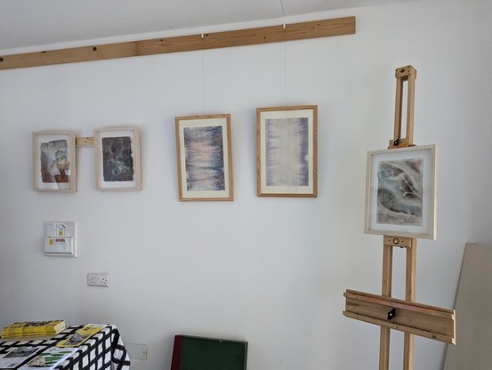 Photo of a gallery wall with 4 abstract paintings hanging in a row and one on an easel in reds and greens. A table with flyers is in front of the paintings