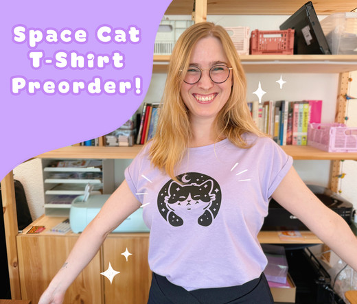 A photo of a girl with blond hair, a bright smile and glasses with her arms out. She’s showing off the tshirt she’s wearing. It’s a lilac purple tshirt with cropped sleeves with a hand printed space cat print on the front in black. In the top left corner it says “Space Cat Tshirt Preorder”