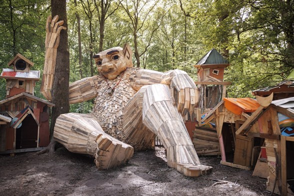 a large wooden figure that resembles a giant lounges on the forest floor, leaning against a tree
