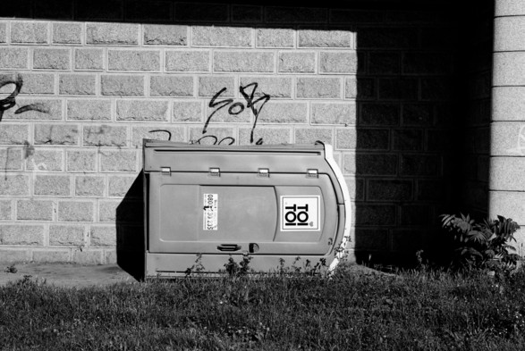 The black-and-white photo depicts a portable toilet lying on its side against a brick wall with graffiti. The wall is partially shaded, and the ground in front of the toilet is grassy with some weeds. The toilet has a label on it with the brand “Toi Toi.”