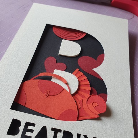 Papercut illustration in progress on a cutting board. A huge letter B with paper elements in it (a bag, a fan, a map, some hearts, some theatre curtains