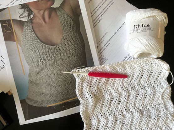 From left to right: a printout of the cover page of the pattern showing a color photograph of a white woman from the chin to top of hip wearing a beige cotton crocheted tank top. The tank top has a bit of a wavy pattern that's worked in one piece from side to side, then the bust, back, and straps are worked in a different smaller pattern that looks like rick-rack.

The photo page is laying on top of left side of the pattern pages. On the right side is a ball of Knit Pick's Dishie yarn in plain white. To the bottom of that, laying atop everything else is the fabric I've created so far by following the pattern (mostly), with a red and white crochet hook through the working loop.