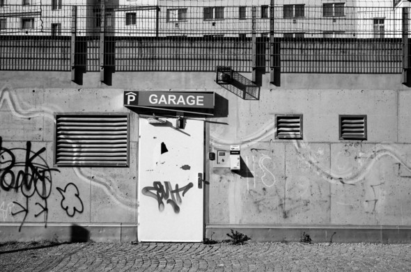 The black and white photo shows a white door with graffiti, labeled “Garage,” set in a concrete wall. The wall also has vents and additional graffiti. Above the wall, a fence and buildings are visible.