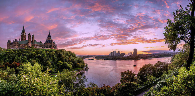 A panoramic of the gothic architecture of the Canadian Parliament Building overlooking the Ottawa River in Ottawa, Ontario, flanked by lush green nature during a magenta sunset after a late-summer day 