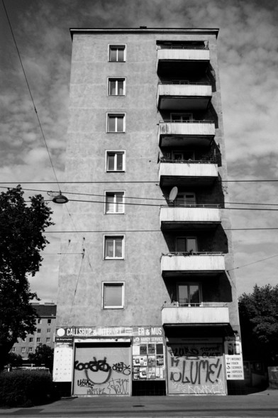 The black and white photo shows a tall, narrow apartment building with a worn exterior. The building has a series of balconies on the right side, and several windows aligned vertically on the left. At street level, there is a storefront with graffiti-covered shutters labeled “CALLSHOP & INTERNET.” Overhead, utility wires cross in front of the building. A tree is visible to the left, adding some greenery to the urban scene.