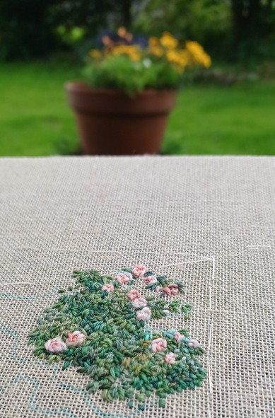 Part worked stitched artwork depicting roses growing on a wall. Behind is an out of focus garden with flowers in a pot and a lawn