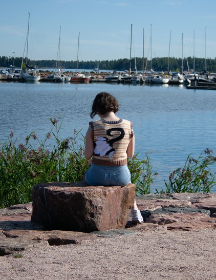 Photo of a woman in a knitted vest. there is water and a lot of boats in the background. There is a picture of a cat on the vest 