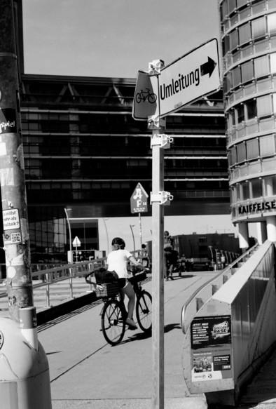 The photo shows a cyclist riding on a path next to a modern building. A sign with the word “Umleitung” (German for “detour”) and a bicycle symbol points to the right. The scene is set in an urban area with contemporary architecture and a clear sky. The cyclist is heading towards a ramp.