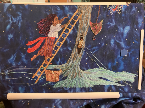 A dark blue starry sky print fabric embroidered with a girl climbing a ladder into a tree with a basket by her feet.  There’s a lantern in the hollow of the tree and its roots are made of water.
