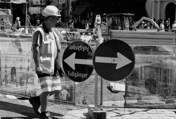 The black and white image shows a construction site with a worker in a hard hat and reflective vest walking past two circular signs. One sign points left and the other right, both labeled “Fußgänger” (pedestrians). In the background, construction equipment and other workers are visible.