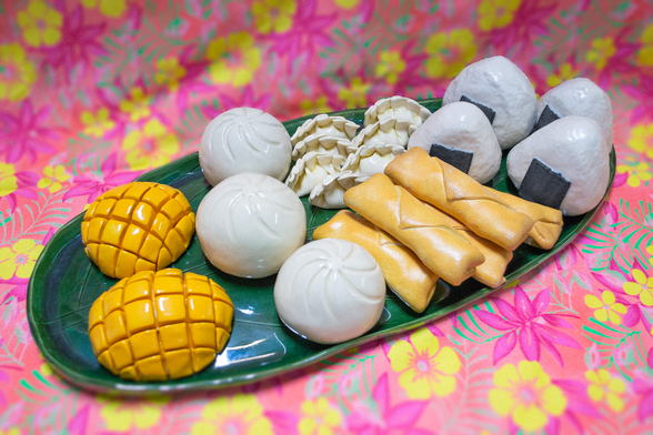 Ceramic onigiri, lumpia, gyoza, siopao, and cut mangos, glazed to look like the real foods. They are arranged on a ceramic banana leaf, which is about 2 feet long. This is all displayed on a pink tropical floral cloth