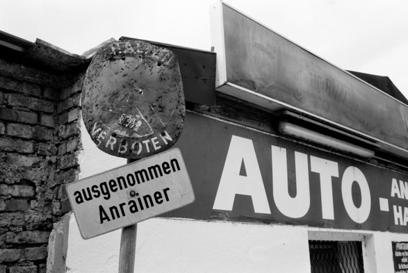 The black and white image shows a weathered and rusty “No parking” sign with the word “Verboten” (forbidden) underneath. Below it, there’s a cleaner sign that reads “ausgenommen Anrainer” (except for residents). Behind the signs, part of a building with a large sign that says “AUTO” is visible. The building’s exterior is a mix of brick and painted surfaces.