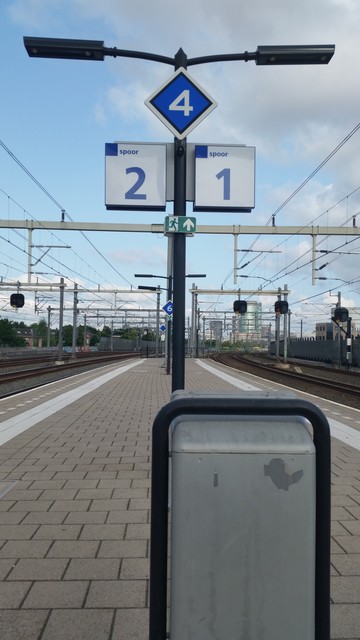 Train platfom grey bin on foreground. Pole with two signs: platform 1 and platfom 2. (blue on white) In the middle a checkered sign with the number 4 in it (white on blue). Overhead wires and train tracks curve to the right. A greenish glass building in the distance.