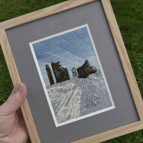 Stitched artwork depicting  ruined mine buildings and the winding wheel in Snow with a blue sky. Framed in an oak frame with grey mount
