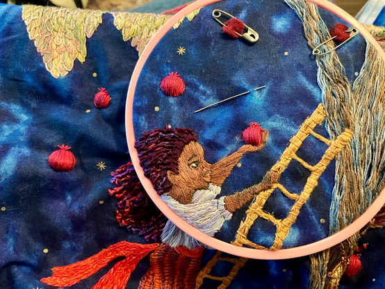 Part of an embroidered image in a small hoop - a girl on a ladder up against a tree holds a pomegranate in her outstretched hand.  Other pomegranates are currently floating in the air around her.